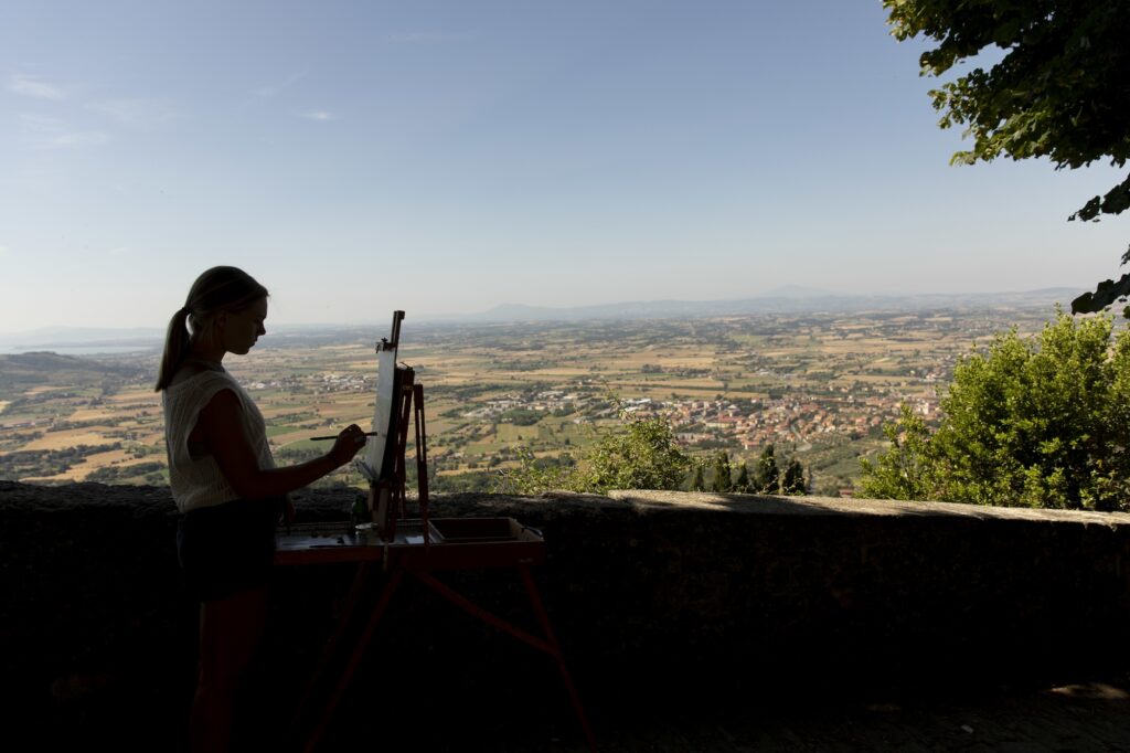 Undergraduate Karley Welch is silhouetted as she paints the Tuscany countryside as she paints outside along the wall below the John D. Kehoe Cortona Center.