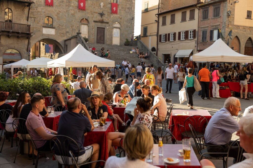 Tourists, local residents, and students enjoy a food and drinks at a local market in the Piazza della Repubblica in Cortona on a sunny afternoon.