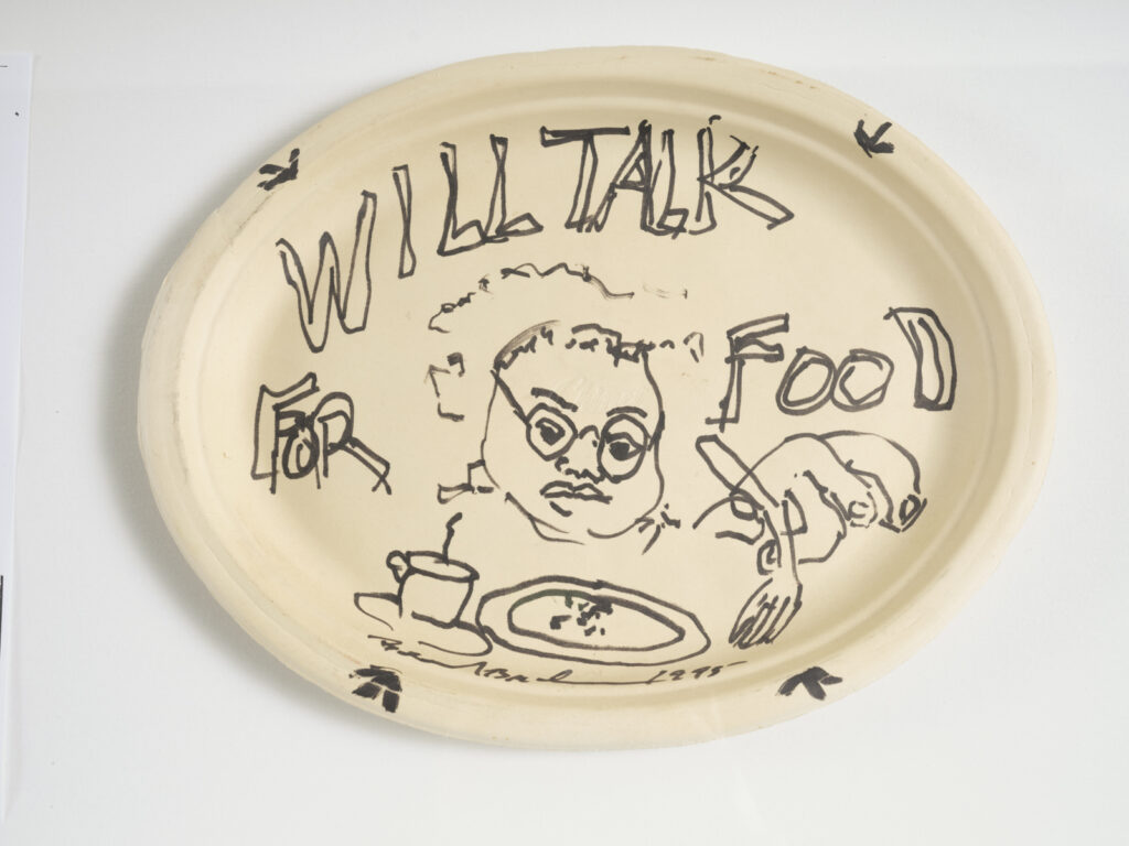Beige paper plate with black marker drawings and graphic text “WILL TALK FOR FOOD” alongside self portrait and hand and fork above plate and mug. Artwork by Beverly Buchanan on view at the Athenaeum for “Beverly's Athens: Beverly Buchanan in Athens, GA”, Spring 2026. Photo courtesy of Walker Bankson.